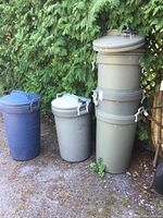 Five Rubbermaid trash cans outdoors. Two gray cans stacked and three individual blue cans with lids and handles.