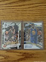 Two Kevin Durant NBA basketball cards side by side on a wooden surface, one with the Brooklyn Nets jersey and one in a Golden State Warriors jersey.