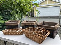 Set of six woven baskets, including three dark brown square baskets, two light brown rectangular baskets with handles, and one shallow light brown open weave basket on a white folding table.