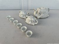 Photo showing two white ceramic teapots with lids and six small ceramic tea cups arranged in a line.