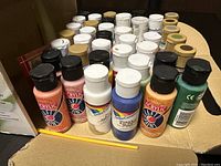 Front and top view of multiple acrylic and craft paint bottles arranged in a cardboard box showing different colored caps and brands