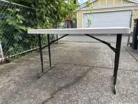 Side angled view showing the foldable table set up outdoors on concrete. White plastic top and black metal legs visible.
