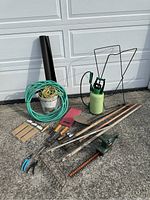 Photo showing garden equipment including hoses, hand trowels, rakes, hoes, sprayer and a long black tube arranged on concrete surface