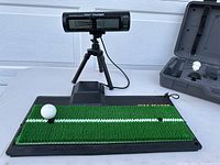 Digital golf swing analyzer device labeled Mitsubishi Electric Golf Trainer on small tripod next to turf hitting mat with a golf ball on tee.
