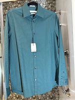Front view of teal Calvin Klein men's dress shirt, long sleeves, with buttons and collar, hanging on a door.