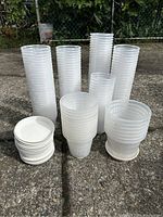 Multiple stacks of clear plastic orchid planter pots of varied heights and diameters with white plastic saucers stacked separately on concrete surface outdoors.