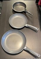 Photo showing three non-stick frying pans arranged vertically on a dark surface, visible from above.