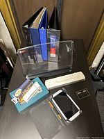 Photo of assorted office supplies including binders, acrylic document holder, laminator, box of clips, calculator, and Samsung tablet with case and stylus on desk.