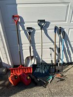 Outdoor gardening tools arranged against a white wall including three shovels, two red plastic jerry cans, a handheld garden rake, and hedge shears.