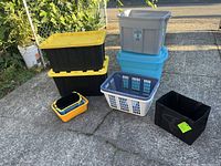Various storage containers arranged outdoors on a paved area, including plastic totes with yellow lids, laundry baskets, and fabric bins.