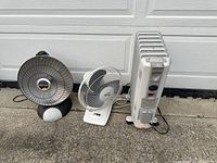 Front view showing the black ceramic space heater, white Hampton Bay desk fan, and white oil-filled radiator heater in a row on concrete outdoors.
