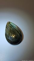 Pear-shaped checkerboard cut Labradorite cabochon showing golden-blue flash, photographed on light background.