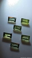 Six rectangular baguette cut green-yellow sapphires arranged on a white surface showing their shape and color variations.