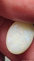 Close-up photo of an oval cabochon natural opal showing play-of-color in blue, purple, and orange hues held between fingers.