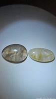 Two natural oval cabochon cut rutilated quartz gemstones shown side by side under light, highlighting golden needle inclusions.