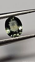 Close-up image showing the natural green sapphire held by tweezers, highlighting its oval cut and color.