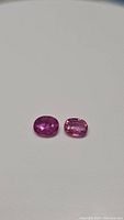 Photo showing two oval cut pink sapphires, different shades of pink, lying separately on a plain surface