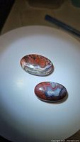 Two oval polished natural red crazy lace agate gemstones shown on a white lit surface highlighting color and patterns.