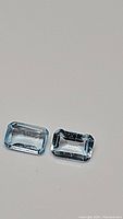 Pair of natural sky blue topaz stones with clear sky blue color and emerald cut, shown laying flat against a plain background.