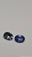 Two oval cut natural tanzanite gemstones with blue to purple coloring shown on plain background.