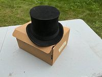 Black top hat placed on original cardboard box, showing full crown and brim from the outside.