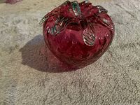 Side angle photo showing the translucent cranberry red glass shaped like a strawberry with textured surface and green glass leaves on top.