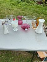 Wide view of glassware collection showing variety of vases, pink bowl, and cups arranged on table outdoors with grass background.