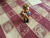 Photo of Hummel porcelain figurine of boy holding teddy bear Christmas ornament on red and beige patterned cloth.