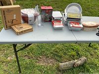 Table with an assortment of kitchen items including disposable aluminum trays, plastic containers, boxed gadgets, and coffee thermos cup set.
