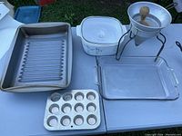 Set of kitchenware including two metal mini muffin trays, white Corning Ware dish, clear glass baking dish, metal juicer with stand and wooden reamer, silver pan, and a cast iron frying pan