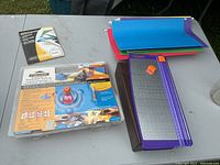 Paper cutter with rotary blade, extra blade pack, and colored laminating pouches on a table outdoors