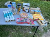Wide shot showing full lot layout on a table outdoors with a cat in the background, displaying shelf liners, assorted candles, paint tools, air diffuser, socket covers, zip ties.