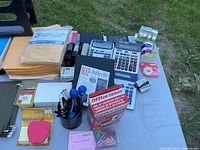 Photo of various office supplies including envelopes, calculators, paper clips, pens, and folders arranged on a table outside.