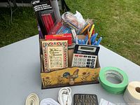 Photo of wooden desk organizer box containing calculator, pens, pencils, a small decorative book, and pack of carpenter pencils.