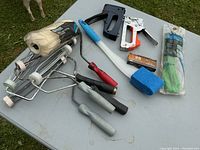 Photo of paint rollers, nail guns, brushes, sponges, lighter, flashlight, and zip ties on gray table outdoors