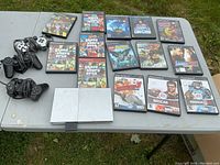 Photo shows a silver PlayStation 2 slim console with visible wear and three controllers: two wired black DualShock and one silver wireless controller. Also displayed are fourteen PlayStation 2 game cases with titles including Grand Theft Auto: San Andreas, Vice City, Raw Danger!, Batman, Madden NFL, NASCAR, and others, laid out on a table outdoors.