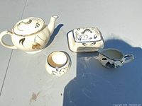 Photo showing all four set items: teapot, butter dish with lid, sugar dish, and cream holder on a white surface under sunlight.