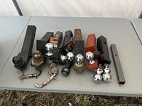 Photo showing an assortment of trailer hitch balls and mounts on a gray surface, including black and red hitch receivers and chrome hitch balls, some with rust and wear.