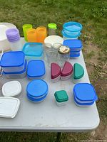 Lot displayed on white table outside with Tupperware containers, glass and plastic plates, plastic utensils, and cups.