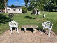 Front view showing two white wrought iron chairs and curved cement bench on patio stones with grass and trees in background.