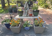 Concrete bench outdoors with various planters and cement statuette arranged on and around it.