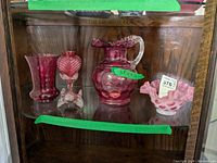 Wide shot showing all six cranberry glass items on a glass shelf inside a wooden cabinet. Contains 4 vases, 1 handled pitcher, and 1 pink fluted bowl.