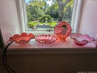 Four cranberry glass items on a window sill: three ruffled bowls of varying sizes and a round footed vase with honeycomb pattern and stopper. All in shades of translucent deep pink-red cranberry glass under sunlight.