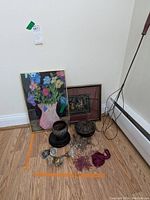 Two framed wall art pieces: a colorful floral painting in a pink vase and a traditional scene, plus two vintage bowls with lids on wooden bases, several beaded tassels, and a metal rug beater with wooden handle placed on floor.