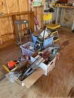Photo showing two chairs (gray kitchen stool and yellow barstool) beside plastic bins and cardboard boxes filled with hand tools inside a wooden garage.