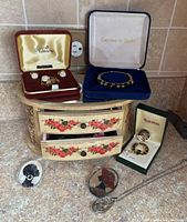 Vintage floral mirror top jewelry box with drawers partially open, surrounded by assorted costume jewelry including boxed sets and loose pins and earrings.