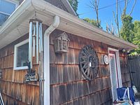 Image showing the corner of a weathered wood exterior wall with a large metal sun face, two round thermometers, wooden insect house, metal sign, and metal wind chimes hanging from the eaves.