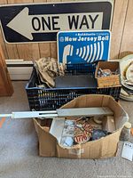 Photo showing 'ONE WAY' sign and 'New Jersey Bell' sign leaning against wood wall, boxed hardware and tags in milk crate and cardboard box with fishing reel.