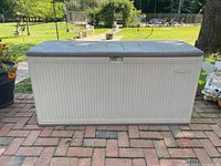 Front view of white Suncast deck box on brick patio with closed gray lid, visible outdoor background.