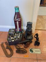 Overview of the lot showing Coca-Cola thermometer shaped like a bottle, box of bottle caps, miniature metal lantern, two small figurines, miniature cannon, and metal hardware tool with Philadelphia Eagles sign on floor